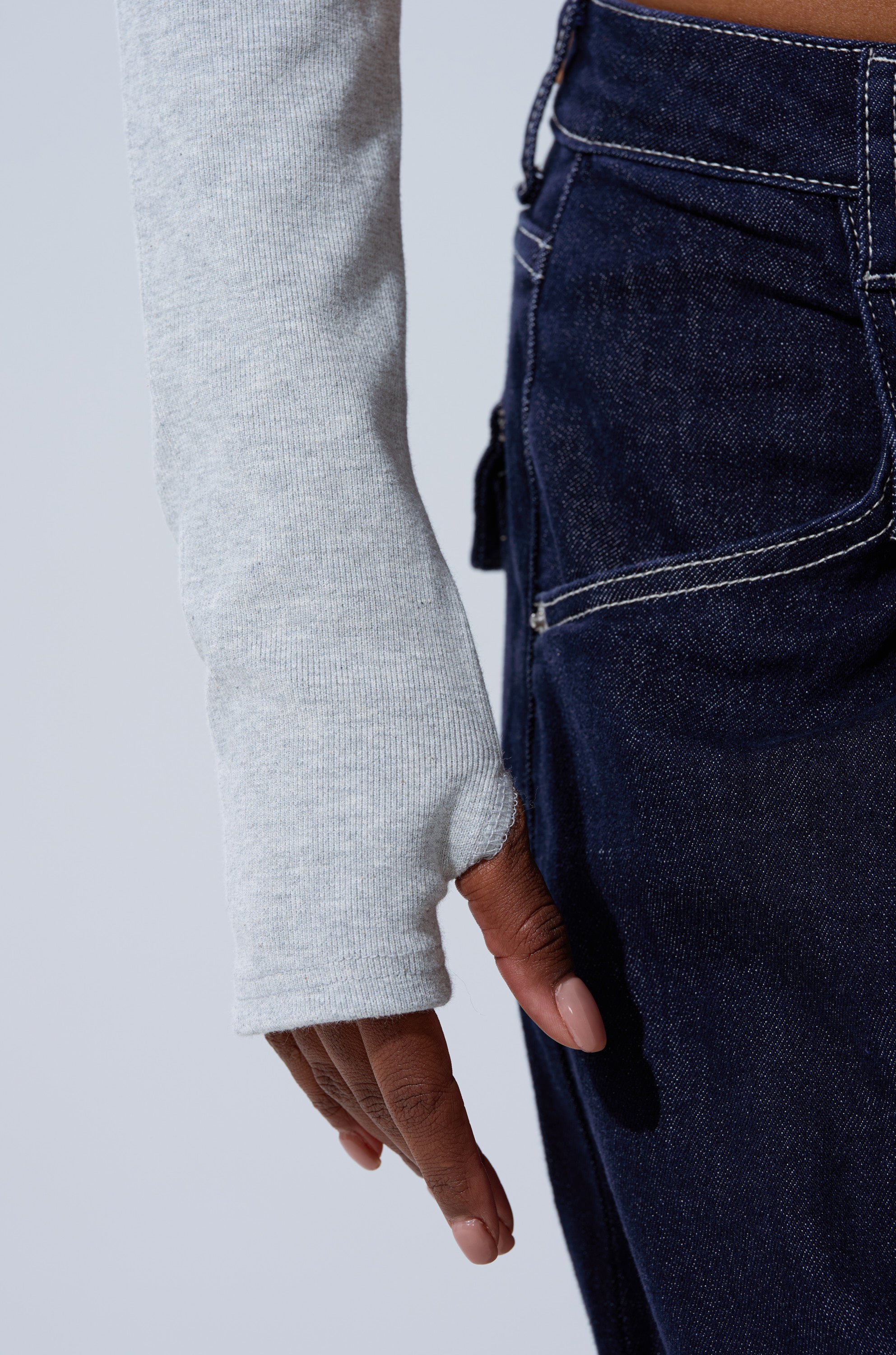 A person wears the BABY GIRL PATCHWORK BOMBER—a cropped Y2K-style jacket—over a light gray thumb-hole long-sleeve shirt and dark blue jeans with white stitching, shown from the waist down with their hand by their side.