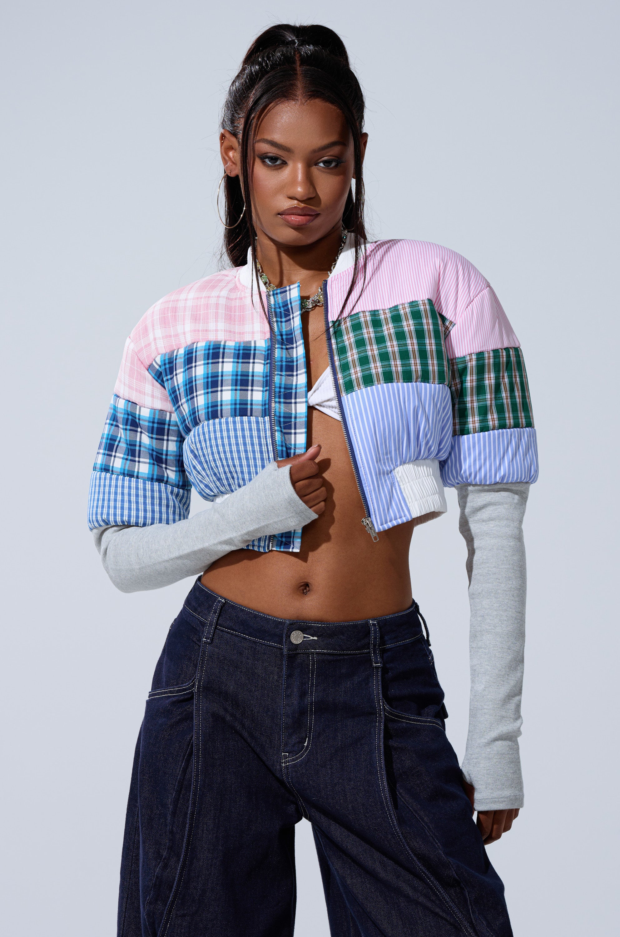 A woman poses confidently in the BABY GIRL PATCHWORK BOMBER, a Y2K-inspired cropped jacket featuring blue, pink, and green plaid and stripes, paired with high-waisted dark denim jeans and gray sleeves.