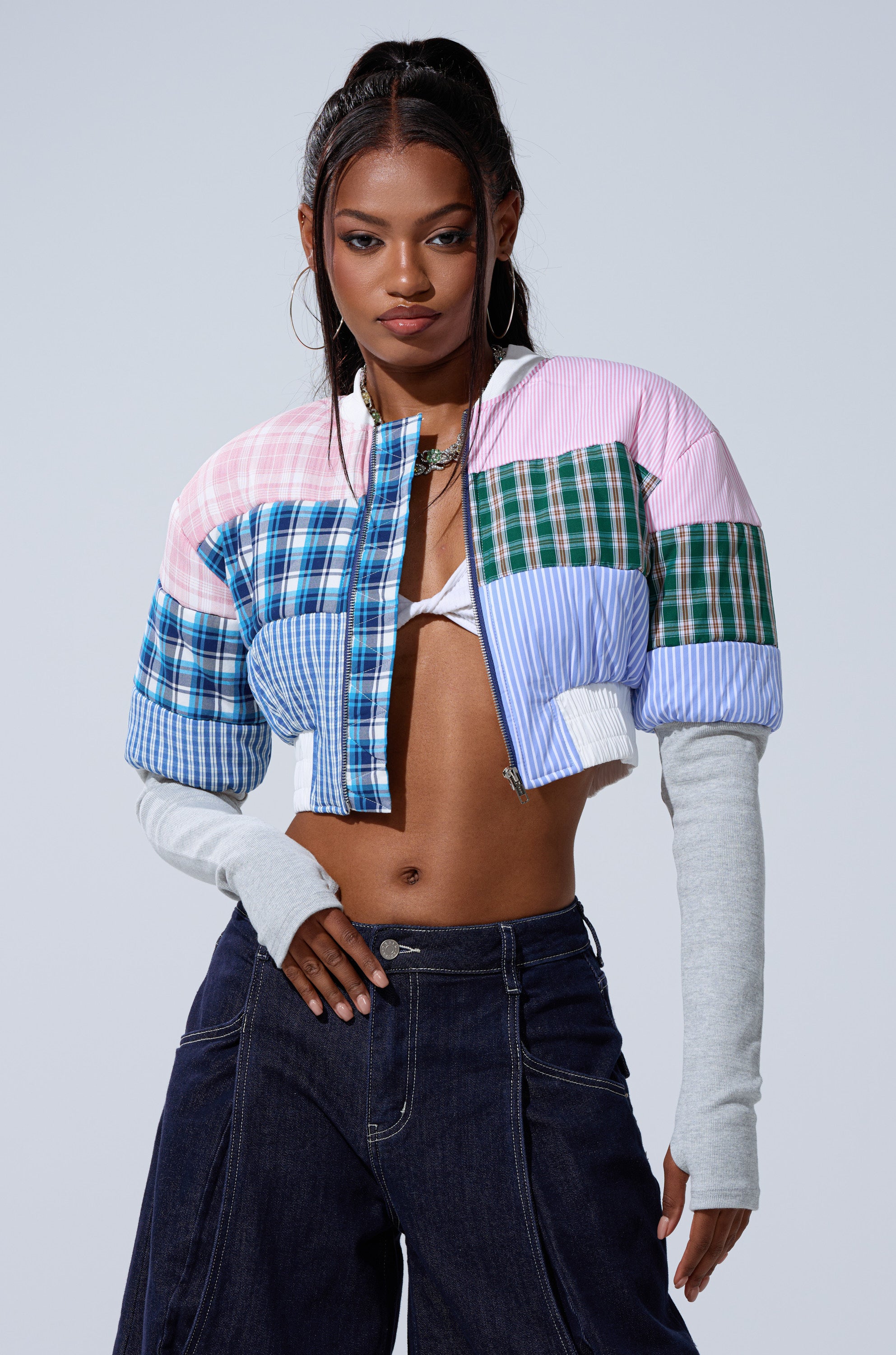 A woman with long dark hair in a high ponytail wears hoop earrings, layered gray sleeves, dark wide-leg jeans, and the BABY GIRL PATCHWORK BOMBER, posing confidently against a plain light background.