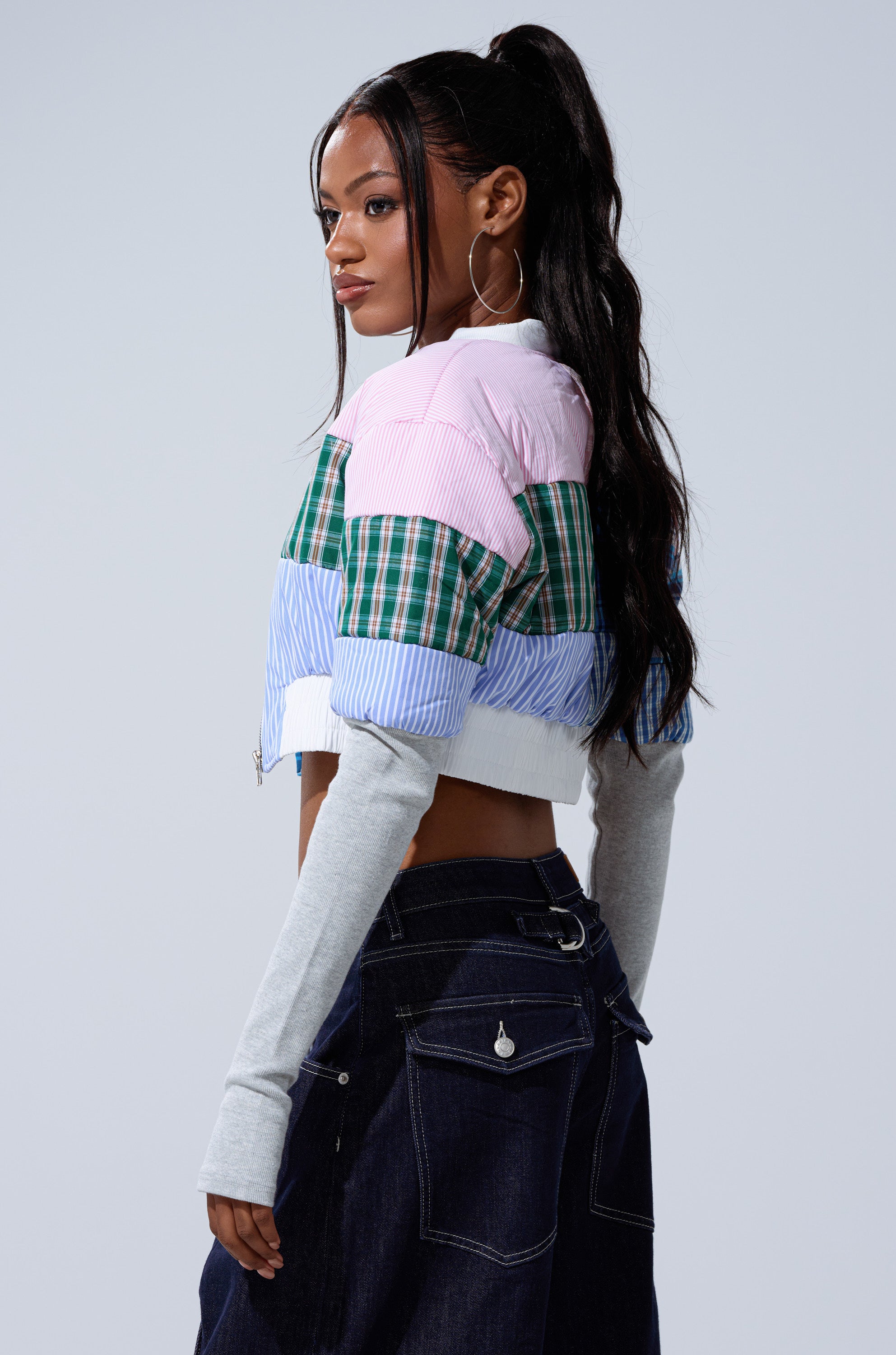 A woman with long dark hair in a high ponytail wears the BABY GIRL PATCHWORK BOMBER and dark denim jeans, standing sideways with a neutral expression against a plain background.
