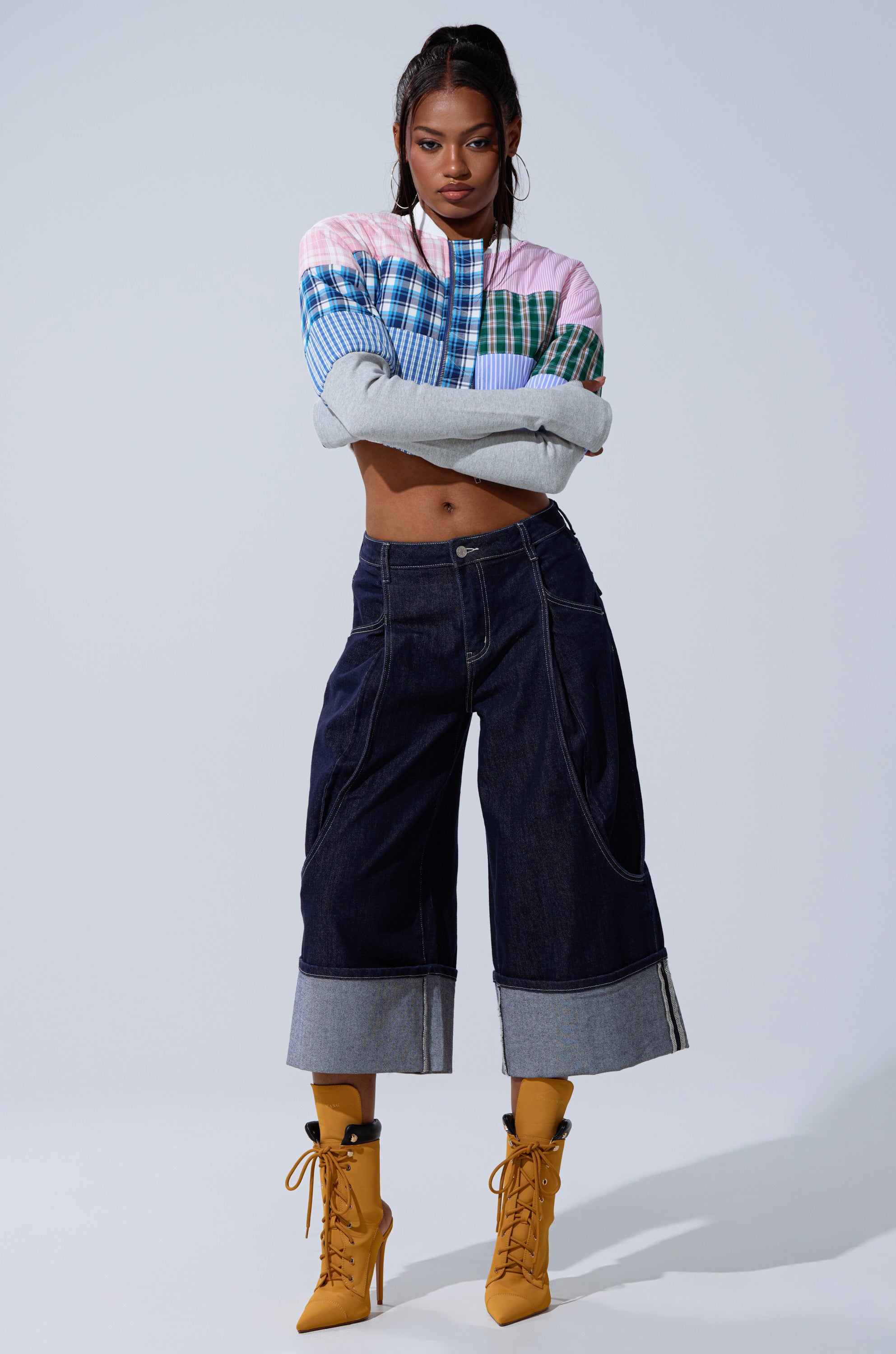 A woman stands confidently against a plain background wearing the BABY GIRL PATCHWORK BOMBER, high-waisted wide-leg dark denim pants with cuffed hems, and mustard-yellow lace-up heeled boots.