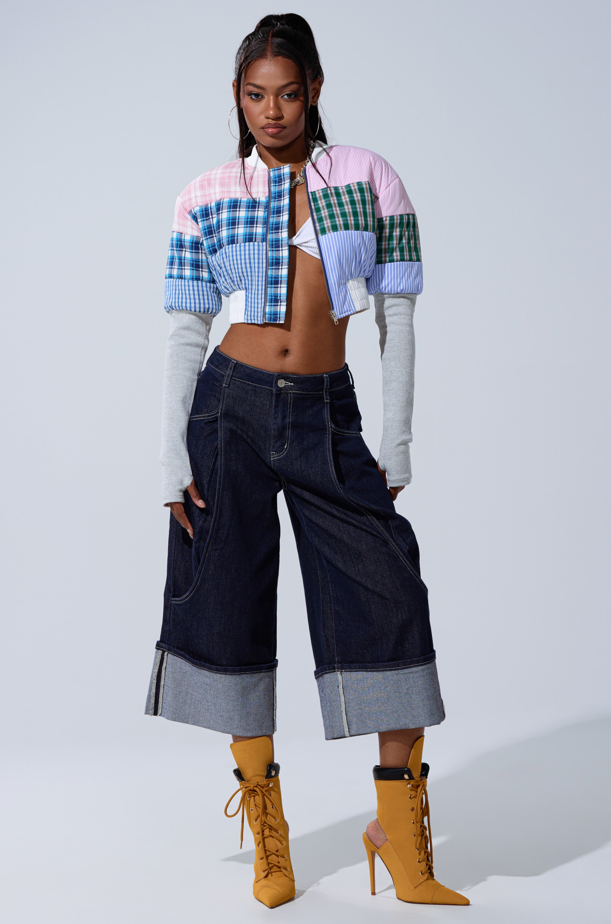 A woman poses confidently in the BABY GIRL PATCHWORK BOMBER, featuring gray sleeves, paired with dark wide-leg cuffed jeans and mustard yellow lace-up heeled boots against a plain background.