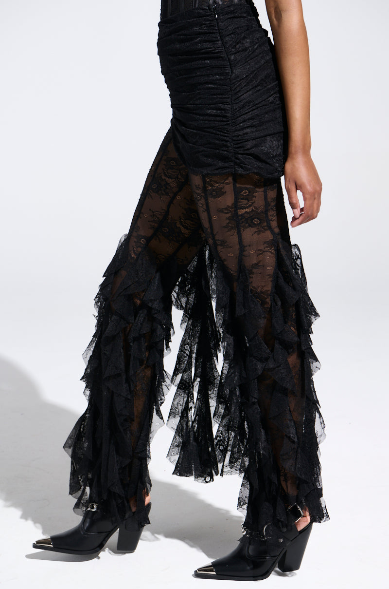 A person wears RUFFLE QUEEN lace skirted leggings with ruffled detailing and black pointed-toe boots. The fitted black top completes the chic, dark outfit against a light background.