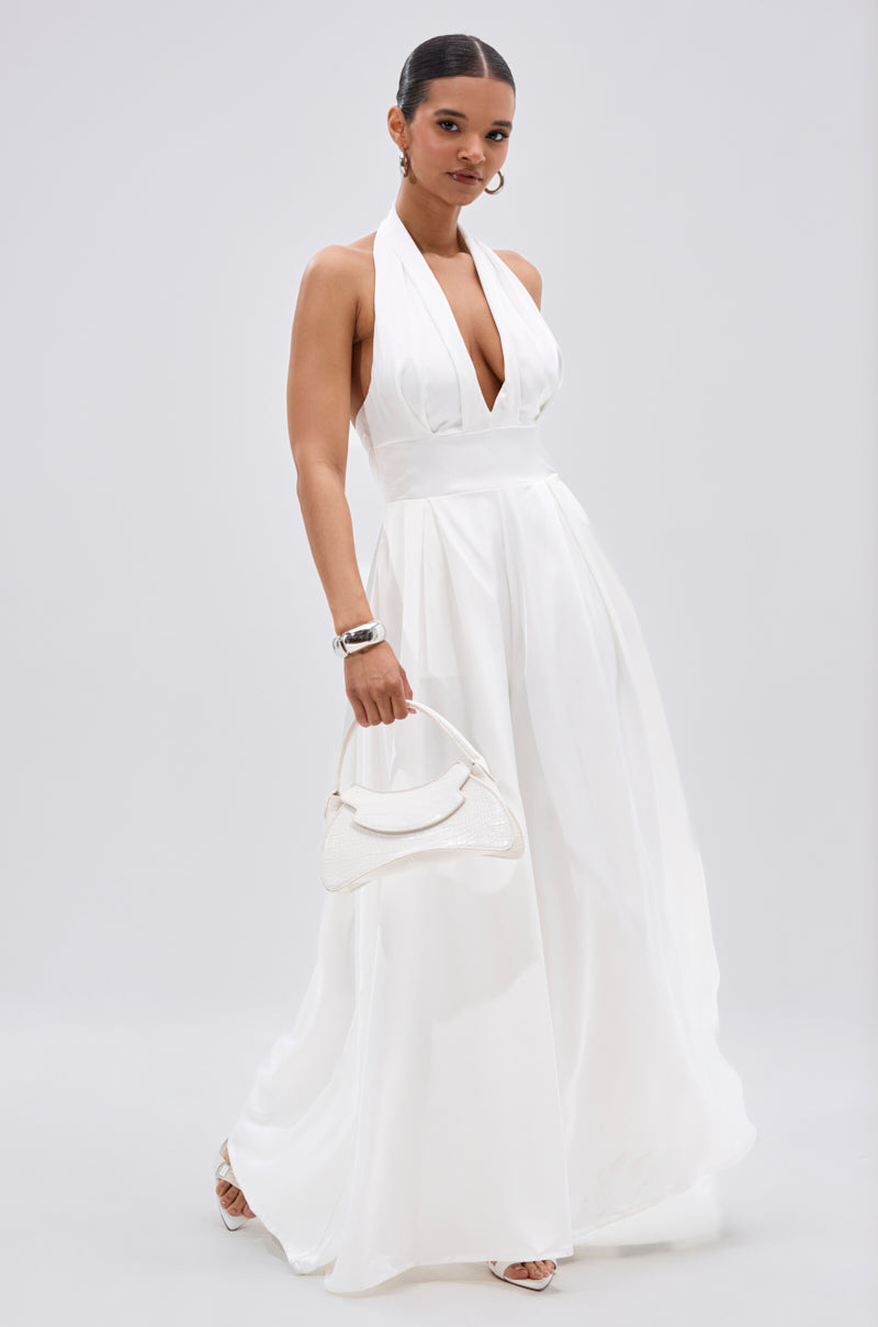 A woman in the UNFORGETTABLE SATIN JUMPSUIT IN WHITE, sleeveless and floor-length with a halter neckline, holds a white handbag. She accessorizes with hoop earrings, a bracelet, and white open-toe heels against a light gray background.