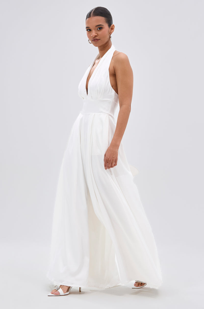 A woman poses against a light grey background in the UNFORGETTABLE SATIN JUMPSUIT IN WHITE, featuring a sleeveless halter neckline and wide-leg pants. She pairs it with silver open-toe heels, hair pulled back for a chic look.