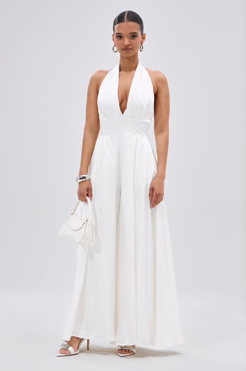 A woman poses against a light backdrop in the UNFORGETTABLE SATIN JUMPSUIT IN WHITE, featuring a sleeveless design, deep V-neck, and wide-leg pants. She accessorizes with hoop earrings, a bracelet, white heeled sandals, and a small white handbag.