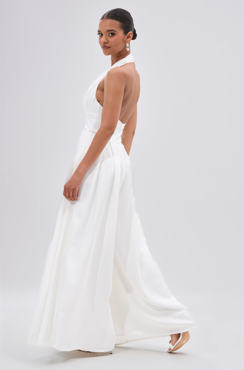 Woman wearing the UNFORGETTABLE SATIN JUMPSUIT IN WHITE, a sleeveless, backless satin jumpsuit with a halter neckline, looking over her shoulder against a plain light background.