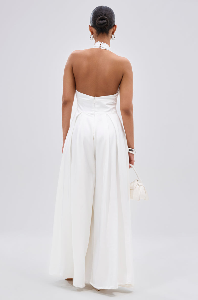 A woman faces away, wearing the UNFORGETTABLE SATIN JUMPSUIT IN WHITE with a halter neckline, wide-leg pants, and open back. She holds a small white handbag and her hair is styled in a neat bun.