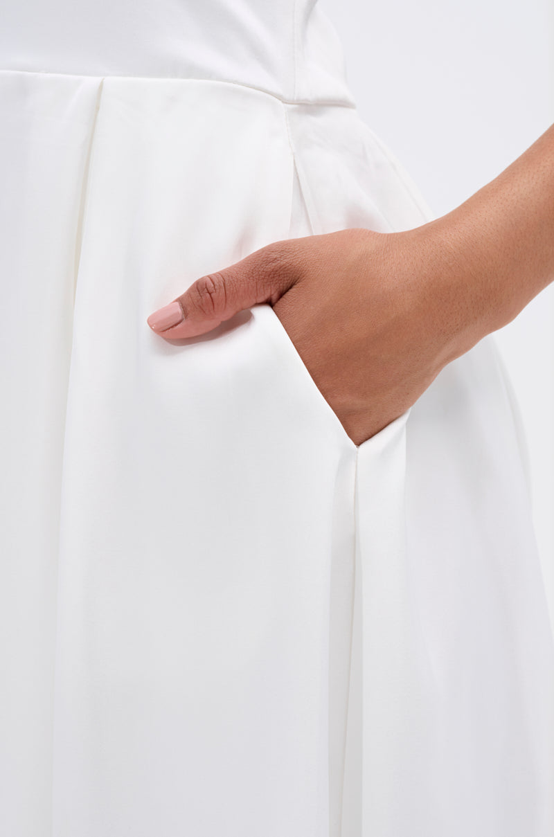 A close-up of a hand with light pink nail polish rests inside the pocket of the UNFORGETTABLE SATIN JUMPSUIT IN WHITE against a plain, light background.
