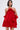 A woman poses against a plain, light background wearing the CHIC AF RUFFLE MINI DRESS IN RED, crafted from lustrous satin with ruffled details, a standout rose embellishment at the neckline, and off-the-shoulder sleeves.