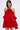 A woman stands against a plain light background wearing the CHIC AF RUFFLE MINI DRESS IN RED, featuring lustrous satin, off-the-shoulder ruffles, and a rose embellishment at the neckline.