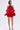 A woman poses against a plain light background in the CHIC AF RUFFLE MINI DRESS IN RED, featuring bold voluminous layers and a rose neckline detail. She has straight dark hair and strappy high-heeled sandals, embodying modern elegance.