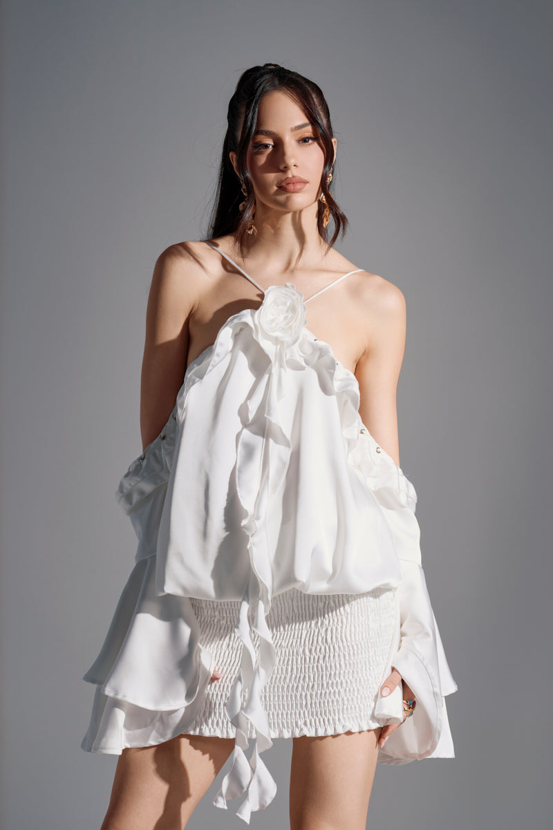 A woman wears the CHIC AF RUFFLE MINI DRESS IN WHITE, featuring ruffled details, a halter neckline, and a large flower accent at the chest, standing against a plain gray background.