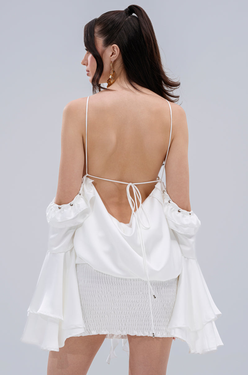 A woman with long dark hair models the CHIC AF RUFFLE MINI DRESS IN WHITE, a backless dress with spaghetti straps and ruffled off-shoulder sleeves, shown from the back against a plain light gray background.
