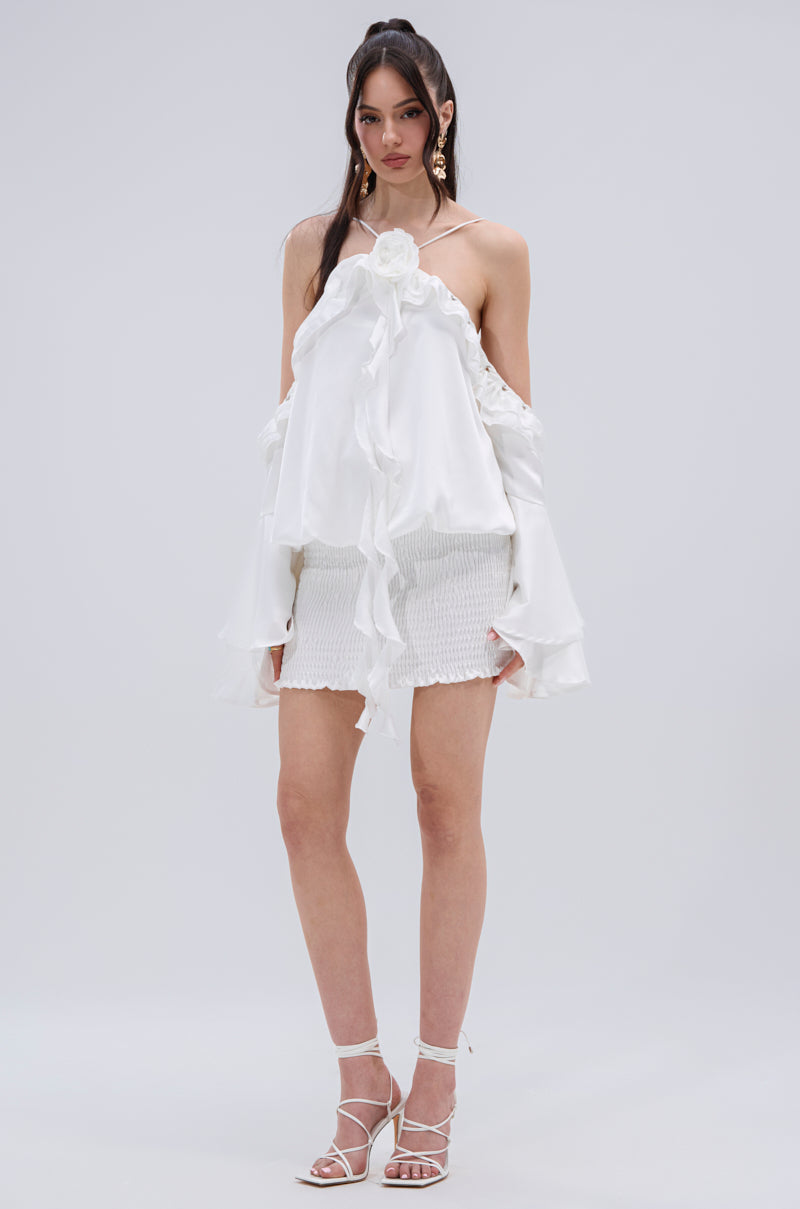 A woman poses against a plain background in the CHIC AF RUFFLE MINI DRESS IN WHITE, featuring high-shine satin, off-shoulder ruffled edges, and a flower detail. She completes the look with strappy heels, statement earrings, and loose hair.