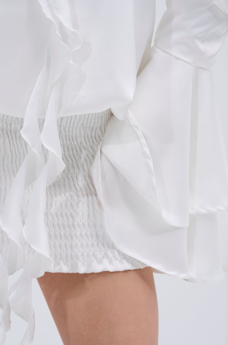 A close-up of a person wearing the CHIC AF RUFFLE MINI DRESS IN WHITE, featuring ruffled sleeves and a white textured skirt. The image focuses on the fabric details and the lower half of the dress.