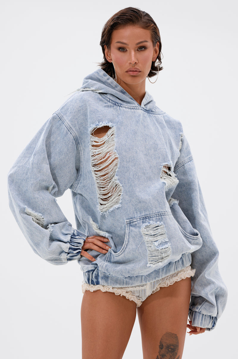 A woman stands confidently in the TRINITY DISTRESSED DENIM HOODIE—an oversized, light-wash piece with bold distressed holes—styled with beige lace-trimmed shorts and hoop earrings against a plain light background.