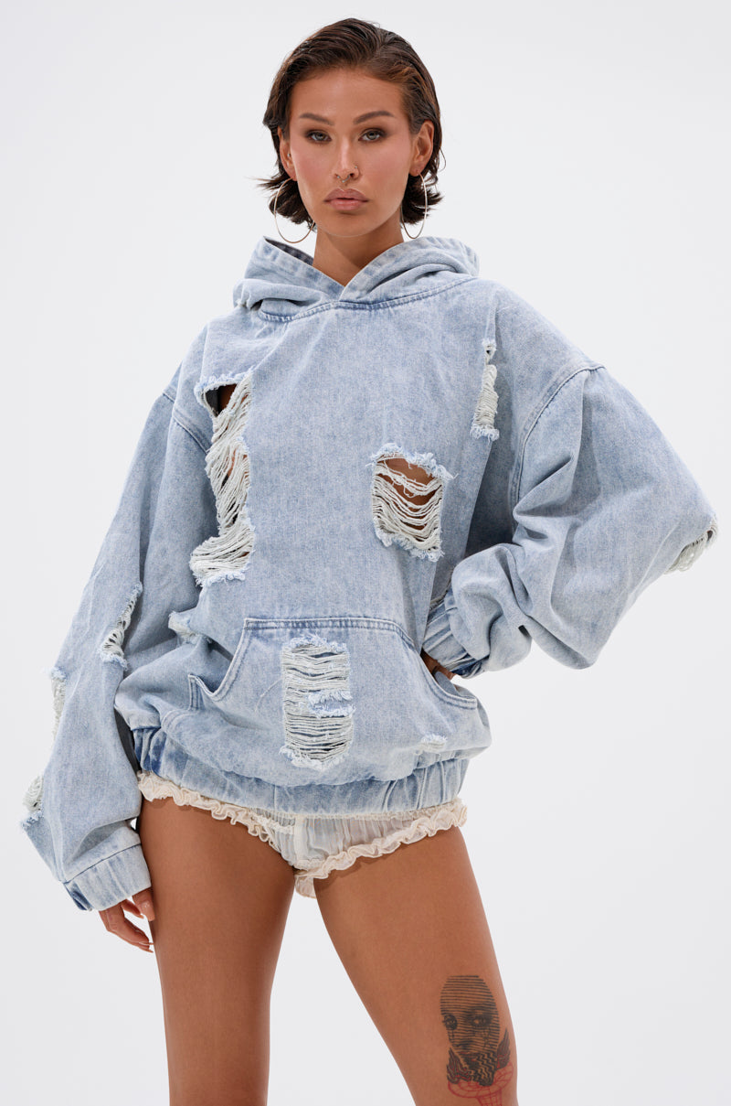 A person poses against a white background in the TRINITY DISTRESSED DENIM HOODIE—an oversized, light-wash denim hoodie with large rips—paired with cream shorts. A tattoo is visible on their left thigh.
