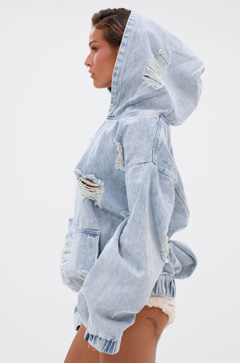 A woman in profile models the TRINITY DISTRESSED DENIM HOODIE—an oversized, light-wash denim hoodie with a large hood and ripped details—paired with lace-trimmed shorts, set against a plain white background.