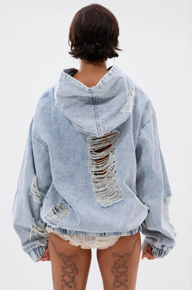 A person with short dark hair, seen from behind, wears the TRINITY DISTRESSED DENIM HOODIE featuring large rips and frayed details, paired with lacy white shorts and lace-up tattoo designs on the backs of both thighs.