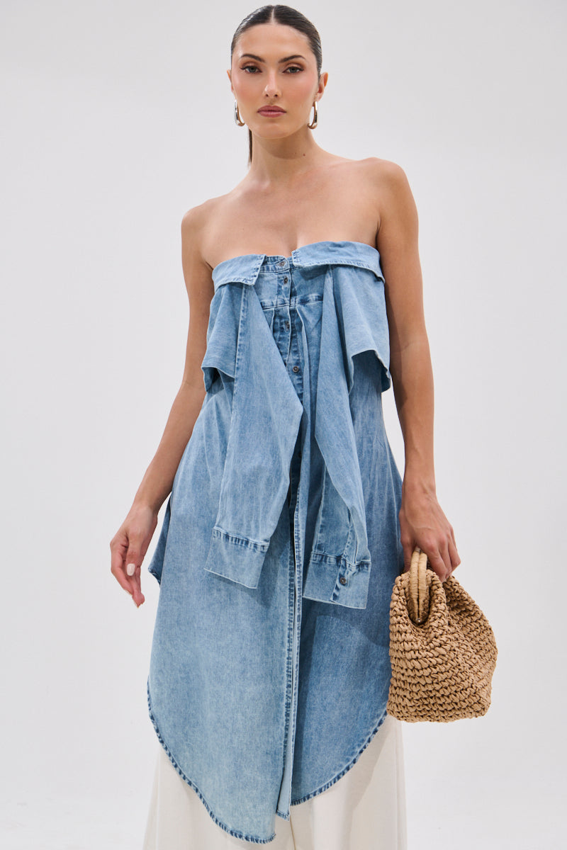 A woman with slicked-back hair and large hoop earrings stands against a white background, holding a woven straw handbag. She wears the CRAZY FOR YOU DENIM BLOUSE, featuring an off-the-shoulder design, draped sleeves, and stonewashed finish.
