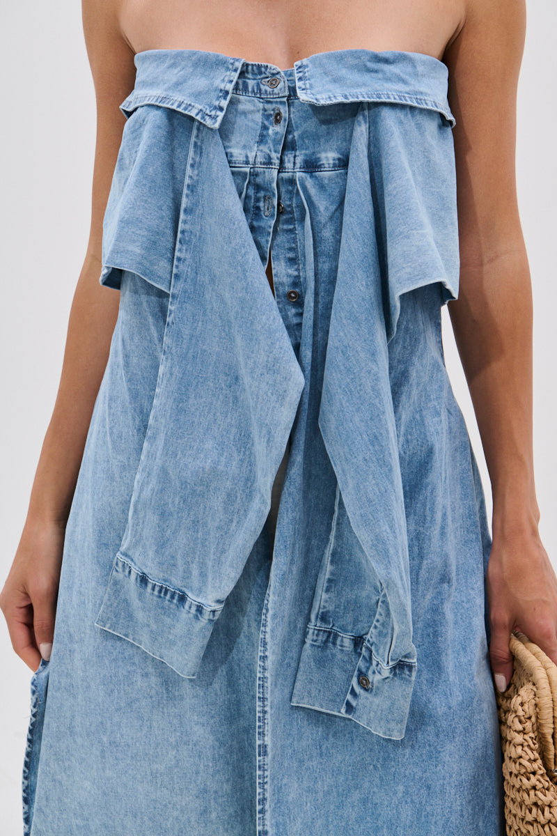 A person wears the CRAZY FOR YOU DENIM BLOUSE, an off-the-shoulder blue denim top with button-down front and layered details, holding a woven straw bag. The image is cropped above the chest and below the waist.