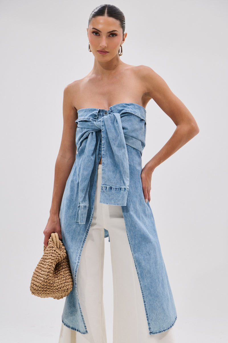 A woman wears the CRAZY FOR YOU DENIM BLOUSE, an off-the-shoulder light blue top with a large front bow and long panels, paired with high-waisted wide-leg white pants and holding a woven straw bag. Her hair is pulled back against a plain background.