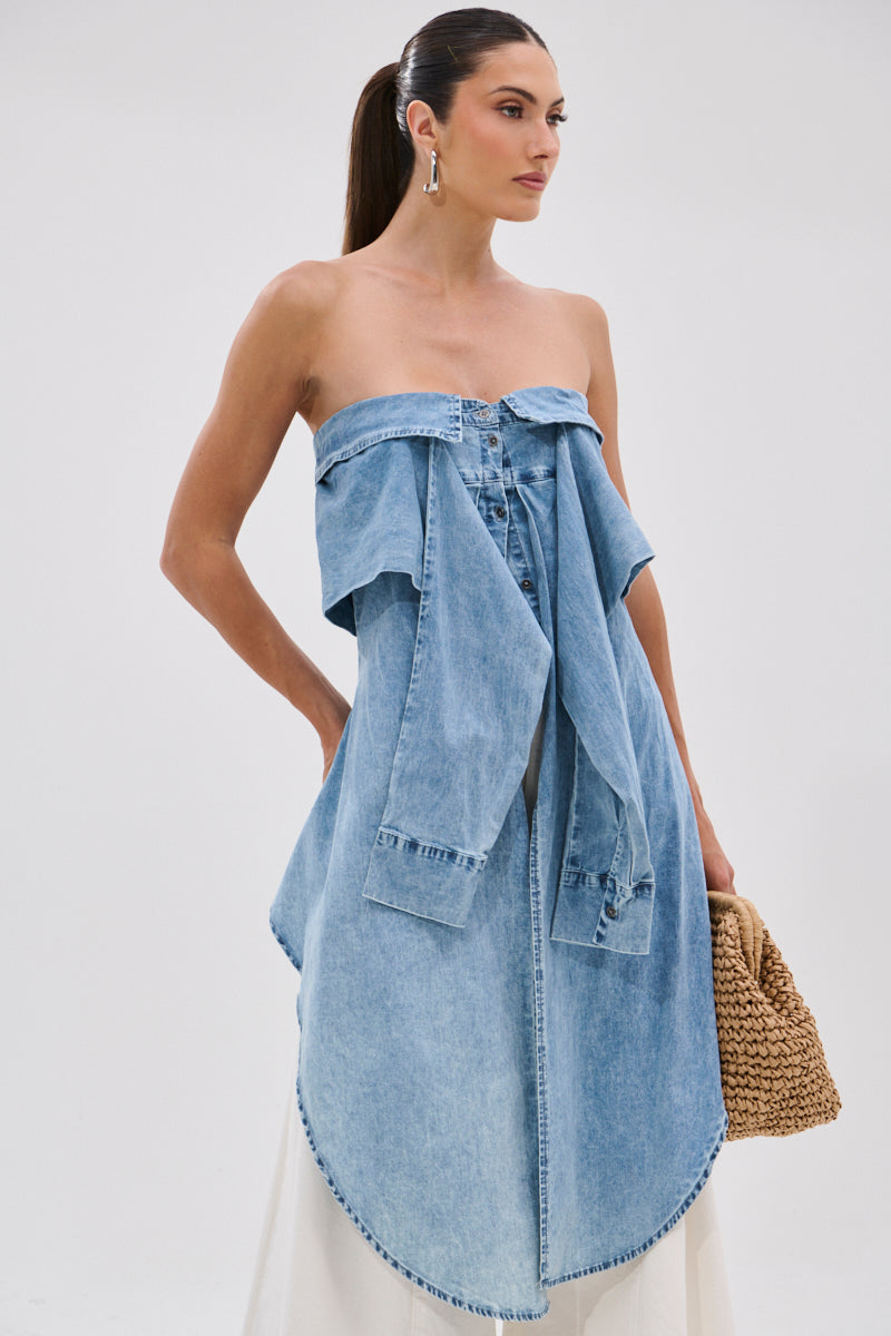 A woman with dark hair in a sleek ponytail wears the CRAZY FOR YOU DENIM BLOUSE styled off-the-shoulder with a stonewashed finish, paired with statement earrings and a woven handbag against a plain background.