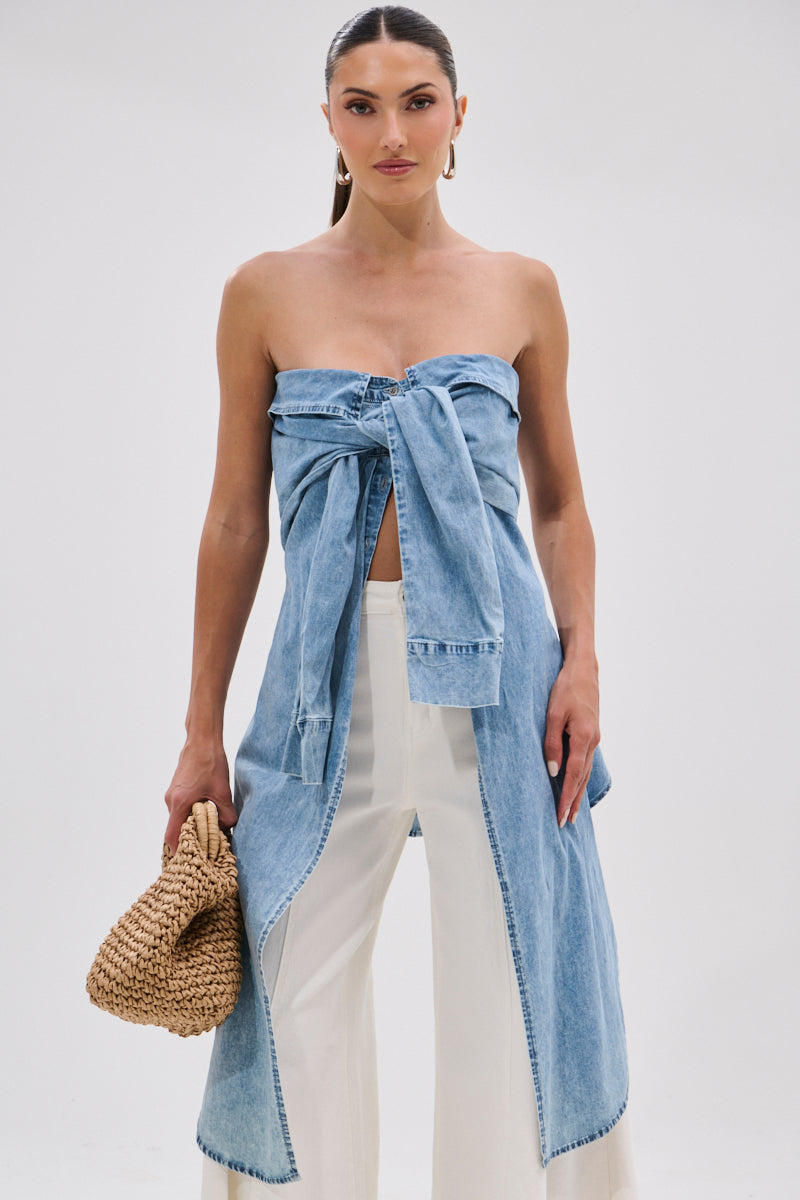 A woman poses against a white background in the CRAZY FOR YOU DENIM BLOUSE, a strapless, off-the-shoulder light blue denim top tied in a large front bow, paired with white wide-leg pants and holding a woven straw clutch.