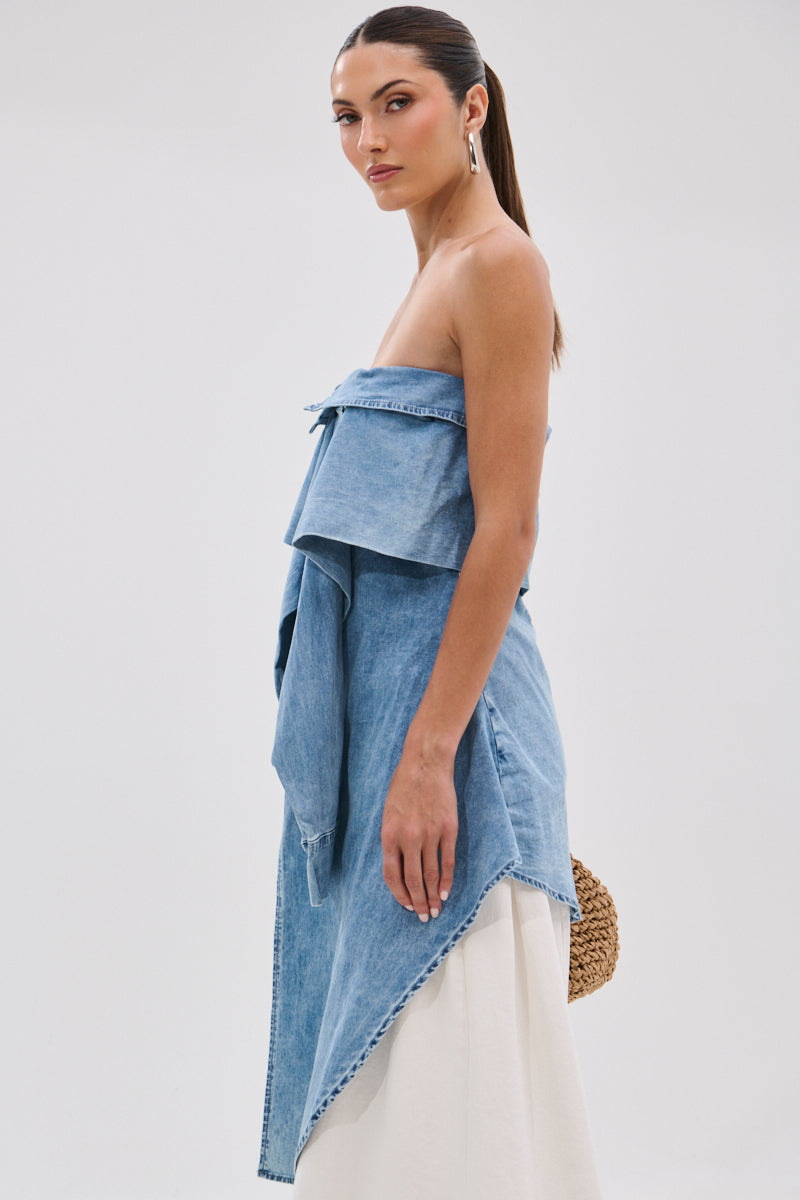 Woman in profile against a plain backdrop wears the CRAZY FOR YOU DENIM BLOUSE—an off-the-shoulder, asymmetrical, stonewashed top—over a long white skirt, hair pulled back, holding a woven round bag.