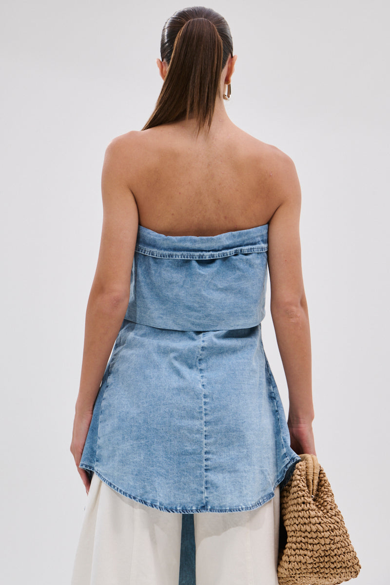 A woman with long hair in a sleek ponytail is seen from behind wearing the CRAZY FOR YOU DENIM BLOUSE, an off-the-shoulder top with a flared hem, styled over a white skirt and carrying a woven bag in her left hand.