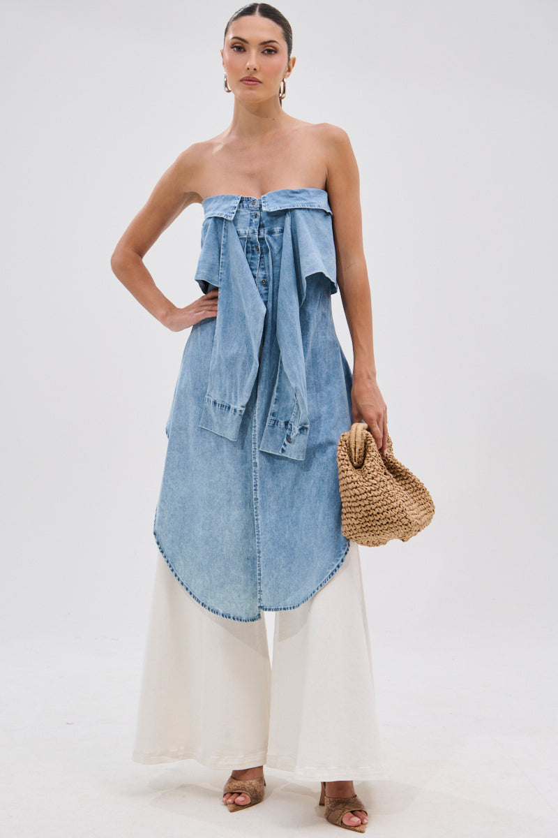 A woman poses against a white background in the CRAZY FOR YOU DENIM BLOUSE—an off-the-shoulder, asymmetric, stonewashed top—paired with wide-leg white pants, open-toe heels, a woven handbag, hoop earrings, and her hair pulled back.