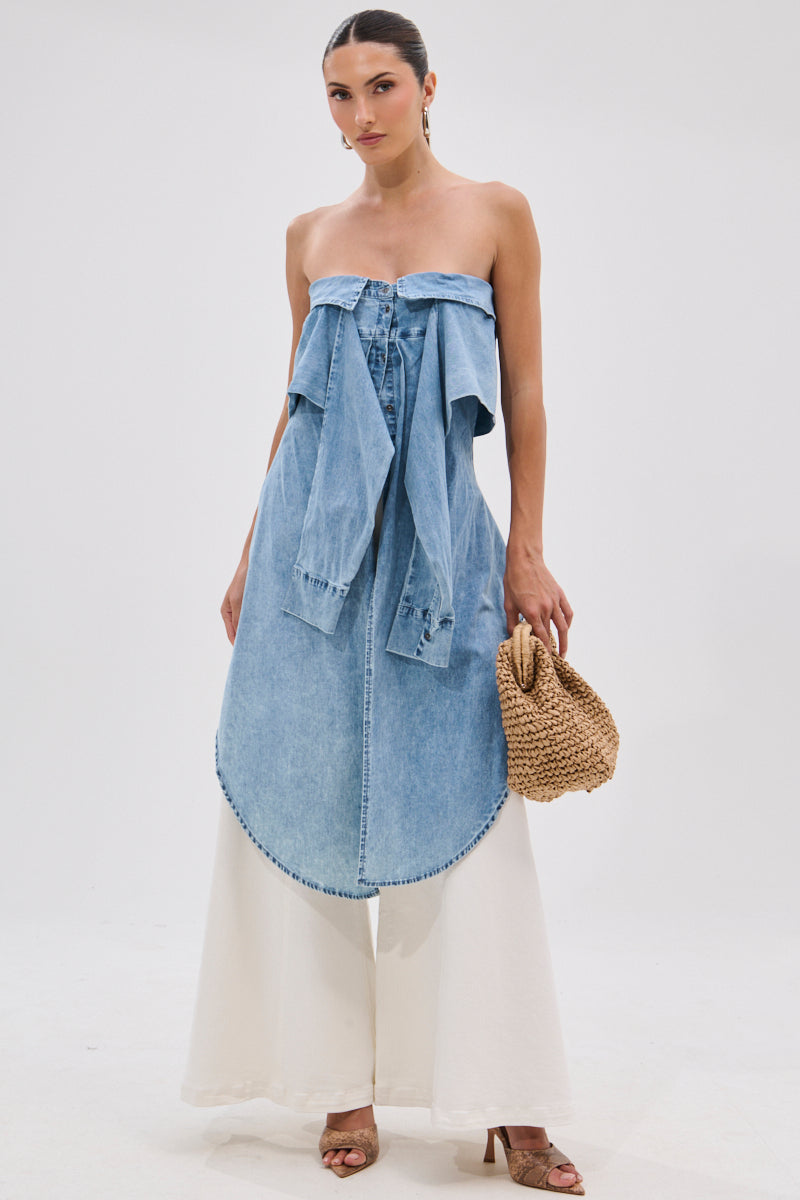 A woman poses in the CRAZY FOR YOU DENIM BLOUSE, styled over wide-leg white pants. She accessorizes with a woven handbag and tan heeled sandals, standing against a plain white background with her hair pulled back.