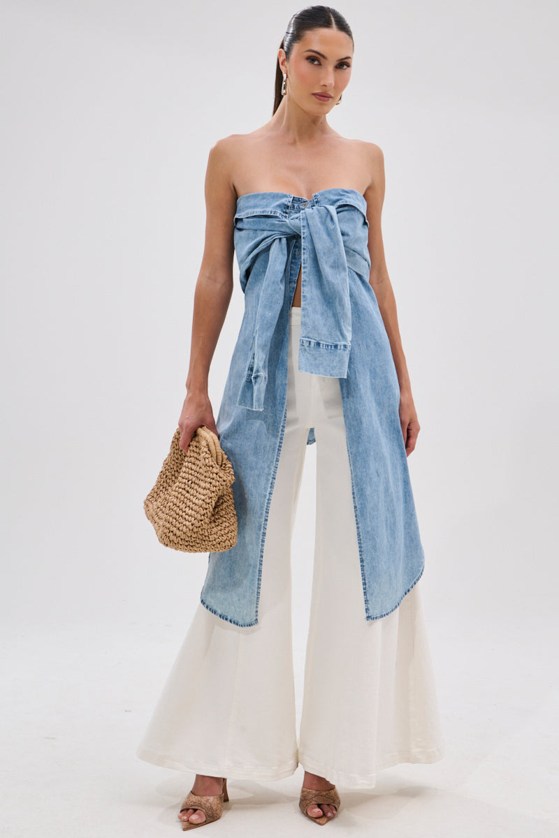 A woman models the CRAZY FOR YOU DENIM BLOUSE in a stonewashed finish, tied at the front and paired with wide-leg white pants. She completes the look with a woven bag, heeled sandals, and a sleek ponytail against a plain background.