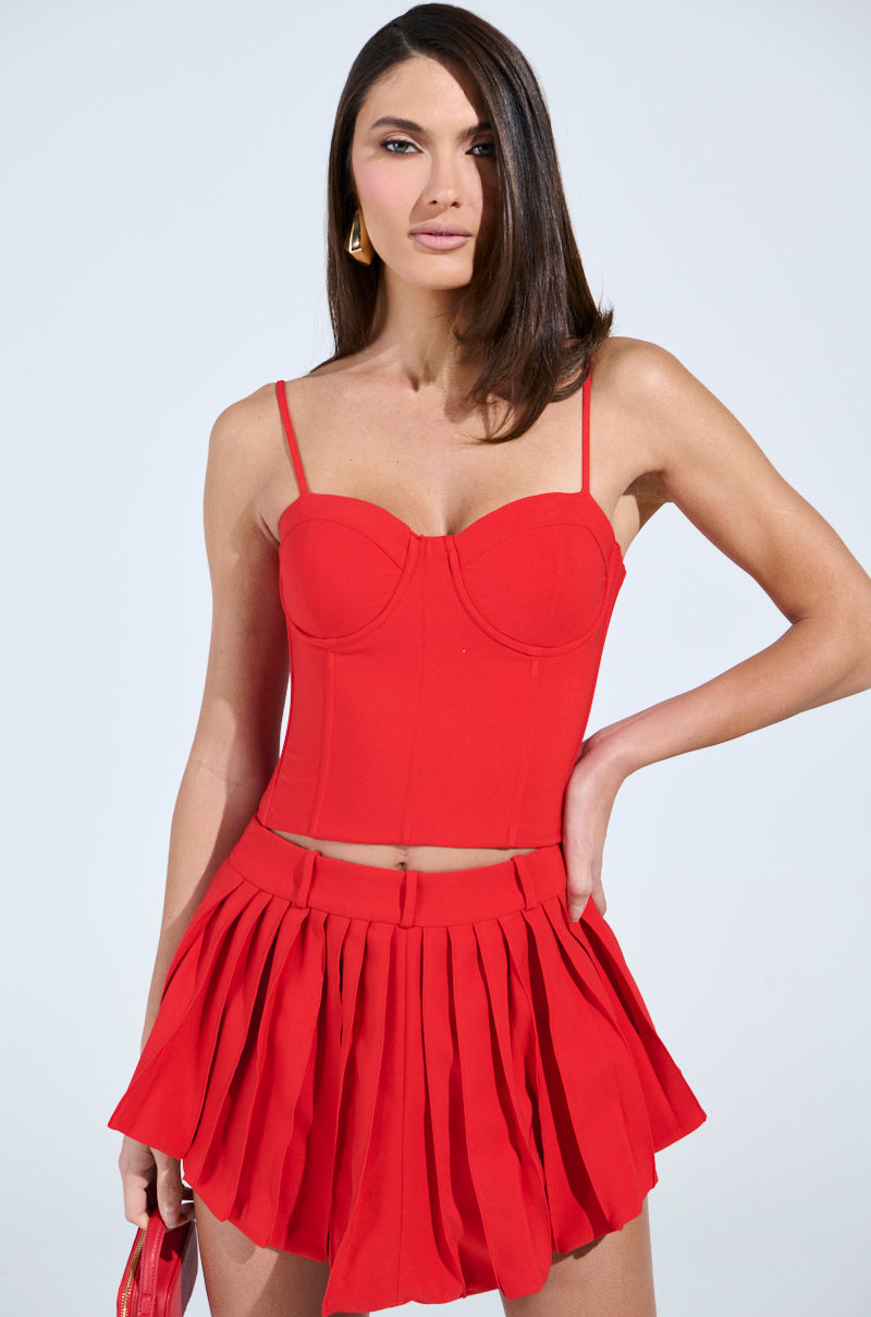 A woman with long brown hair wears the stylish LADY LUX CORSET spaghetti strap top and matching pleated skirt in vibrant red, featuring a sweetheart neckline. She completes her look with a chic red purse against a plain, light background.