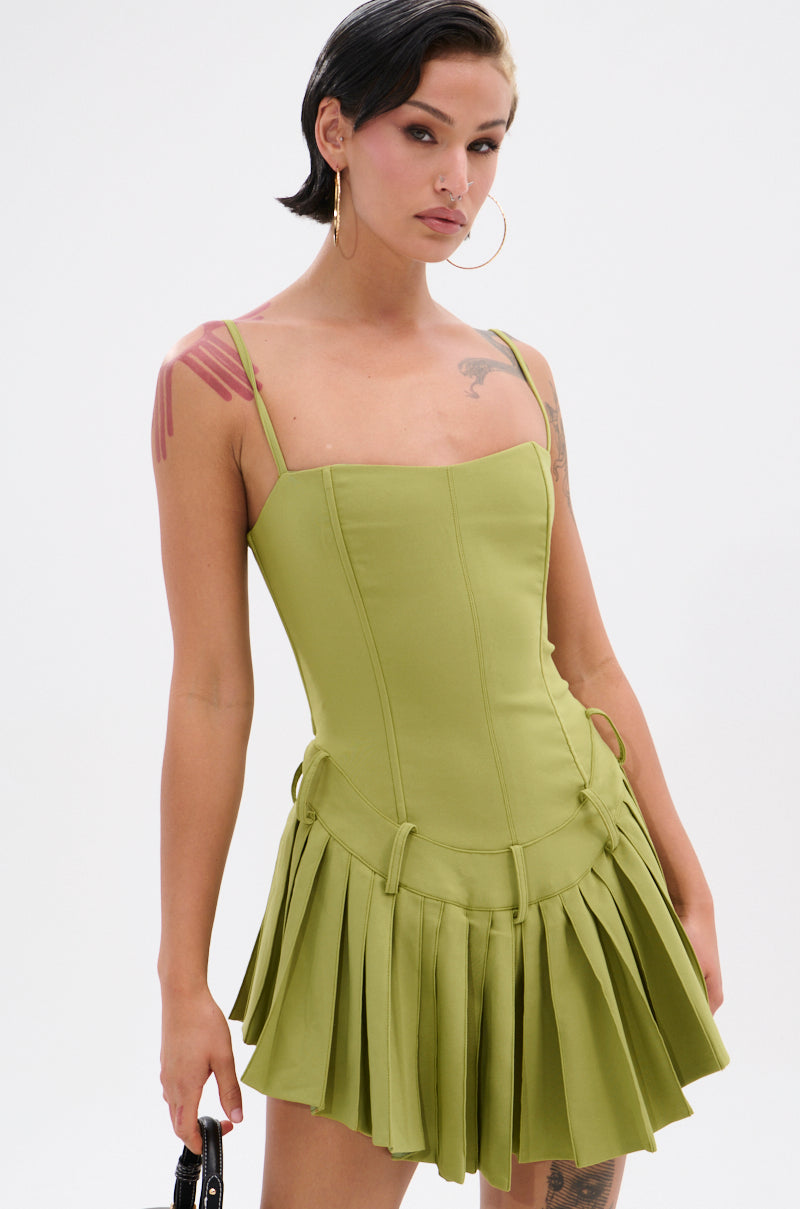 A woman with short dark hair and hoop earrings wears the DANCING THROUGH LIFE MINI DRESS IN OLIVE, a fitted mini with thin straps and a pleated skirt. She has tattoos, holds a small black bag, and stands against a plain white background.