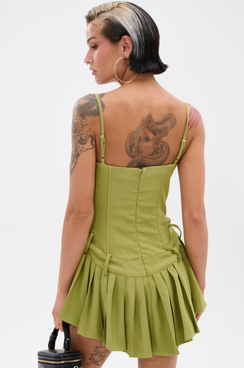 A woman with short, dark hair and a blonde streak wears the DANCING THROUGH LIFE MINI DRESS IN OLIVE, featuring a corset-style bodice and thin straps. Tattoos on her arm and back are visible as she faces away, holding a black bag.