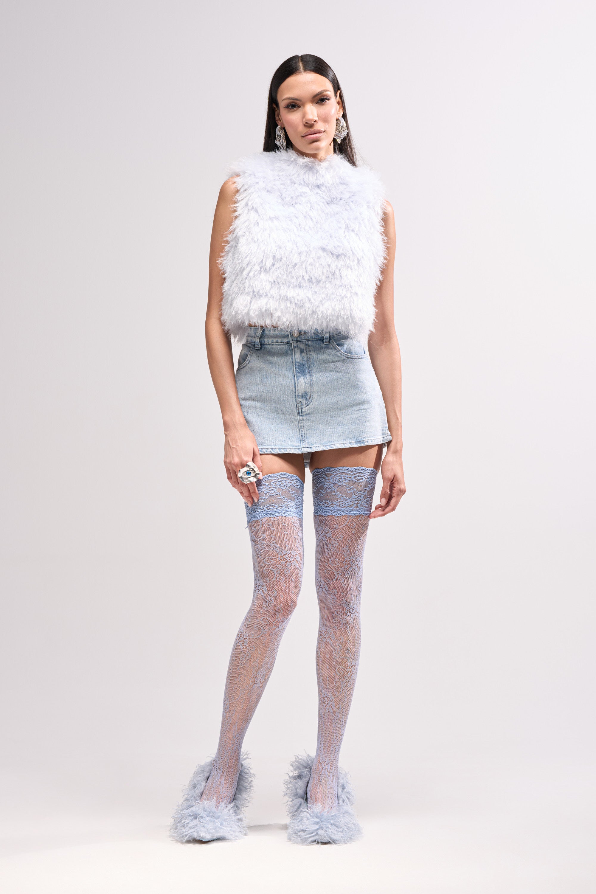 A woman stands against a plain background wearing a fluffy light gray sleeveless top, the WAYS TO GO TIE DENIM SKIRT, sheer blue lace thigh-high stockings, fluffy matching shoes, and large dangling earrings.