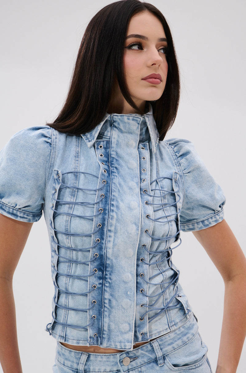 A woman with long dark hair wears the WAYS TO GO TIE DENIM BLOUSE, a light blue top with puff sleeves, lace-up detailing, and silver eyelets. She stands slightly turned right against a plain, light background.