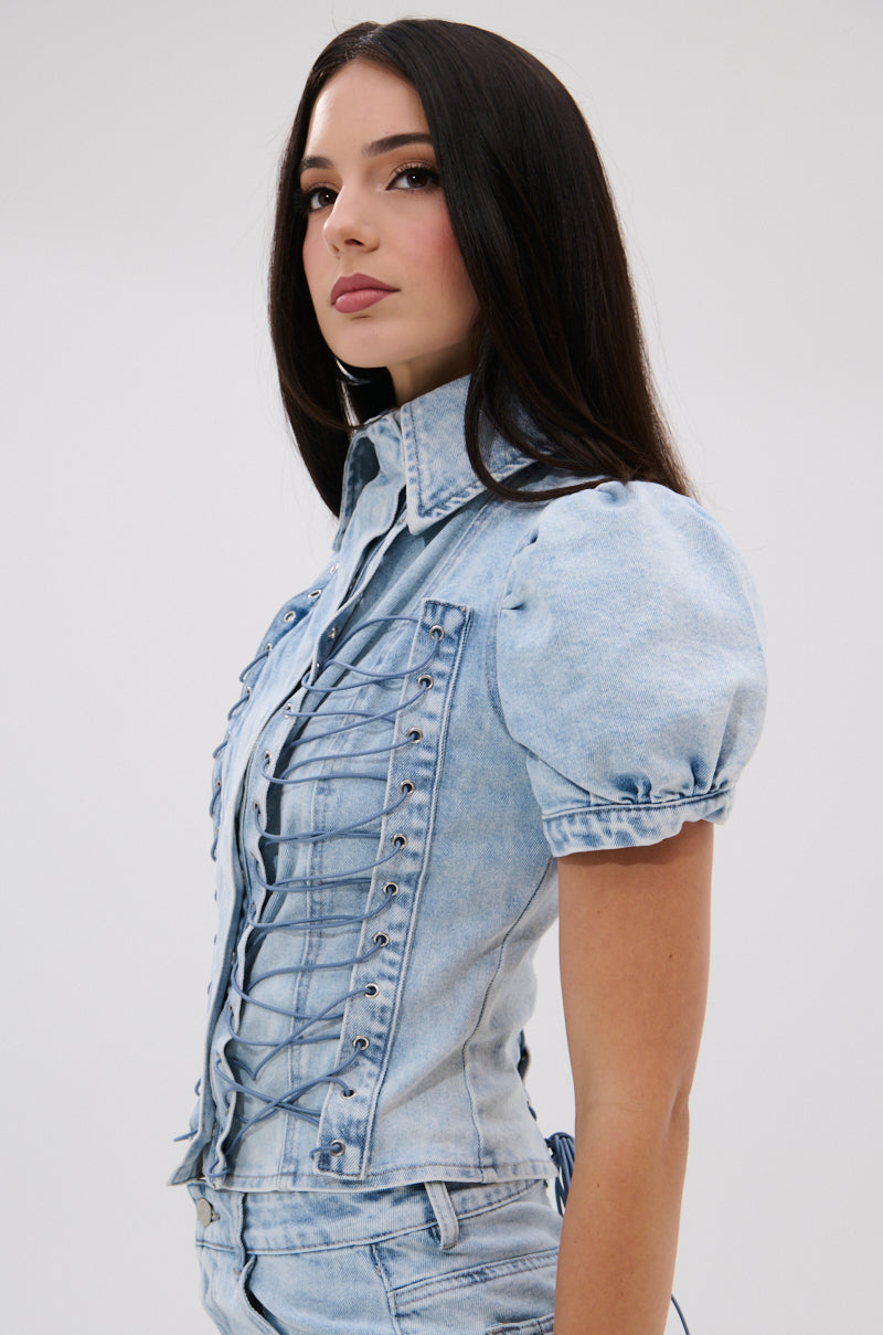 A woman with long dark hair models the WAYS TO GO TIE DENIM BLOUSE, which features short puff sleeves and a lace-up front. She is pictured in profile against a plain light background, looking confidently ahead.