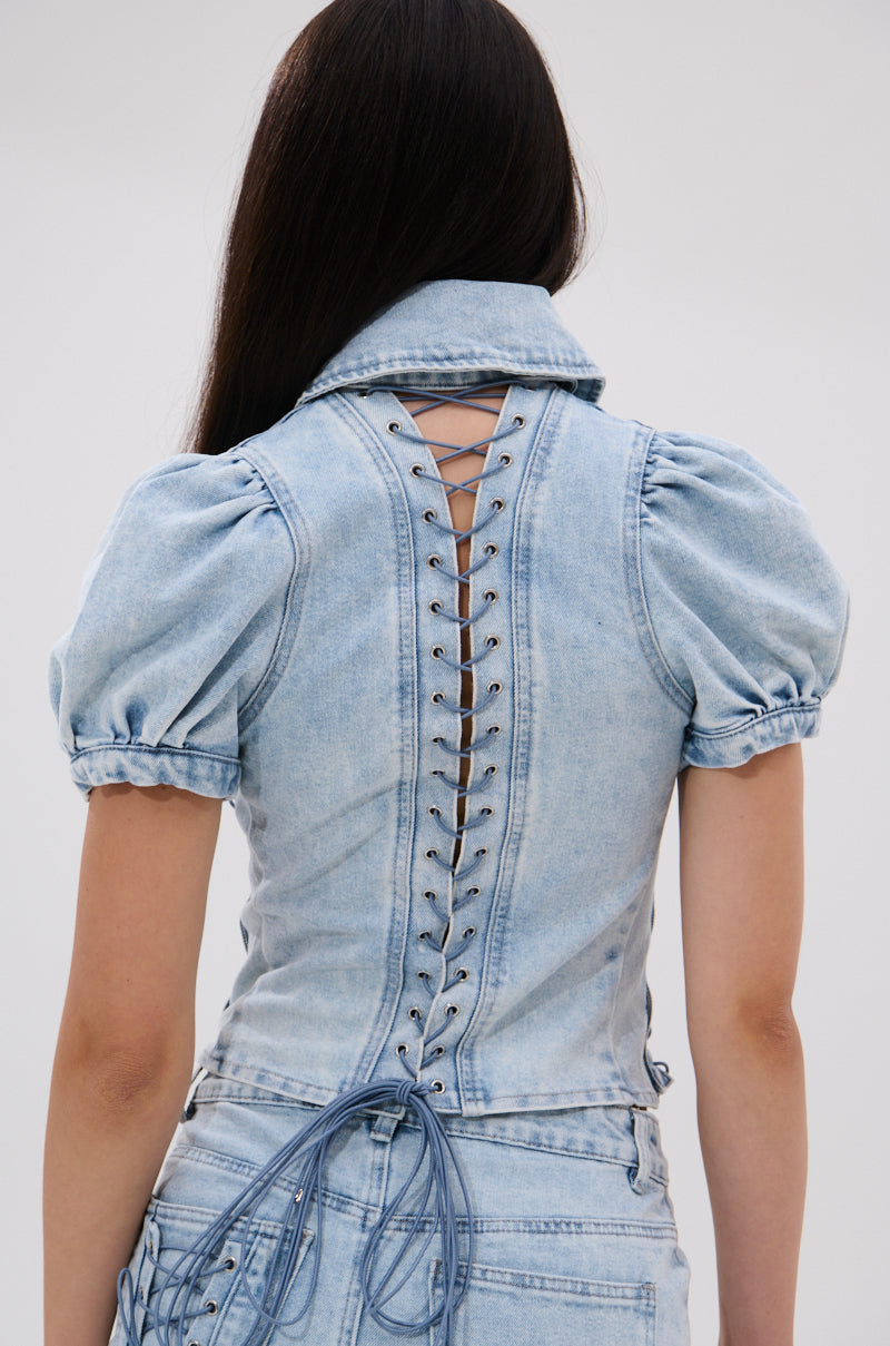 A woman with long dark hair is seen from the back wearing the WAYS TO GO TIE DENIM BLOUSE, a light blue puff sleeve top with lace-up eyelet details down the back, paired with matching denim bottoms for a coordinated look.