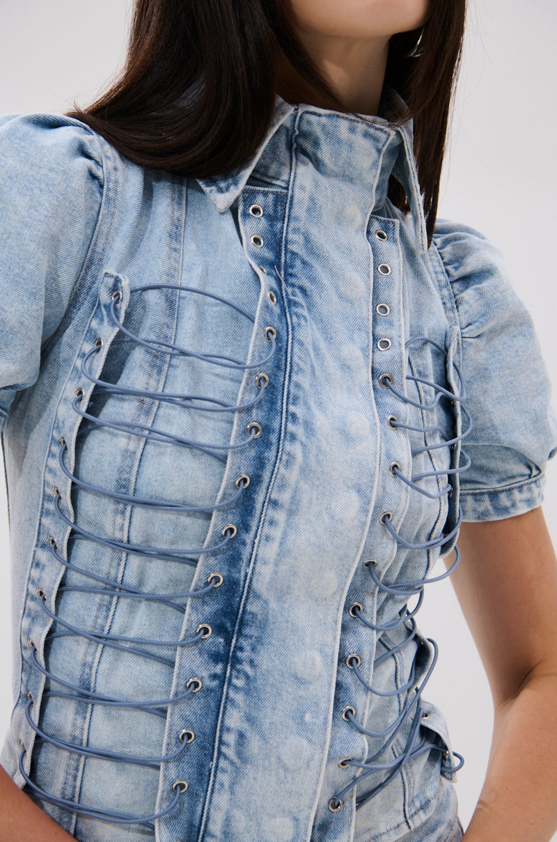 A woman wears the WAYS TO GO TIE DENIM BLOUSE, a light blue acid-wash top featuring puffed short sleeves and vertical decorative lacing on the front. The photo is cropped from shoulders to waist.