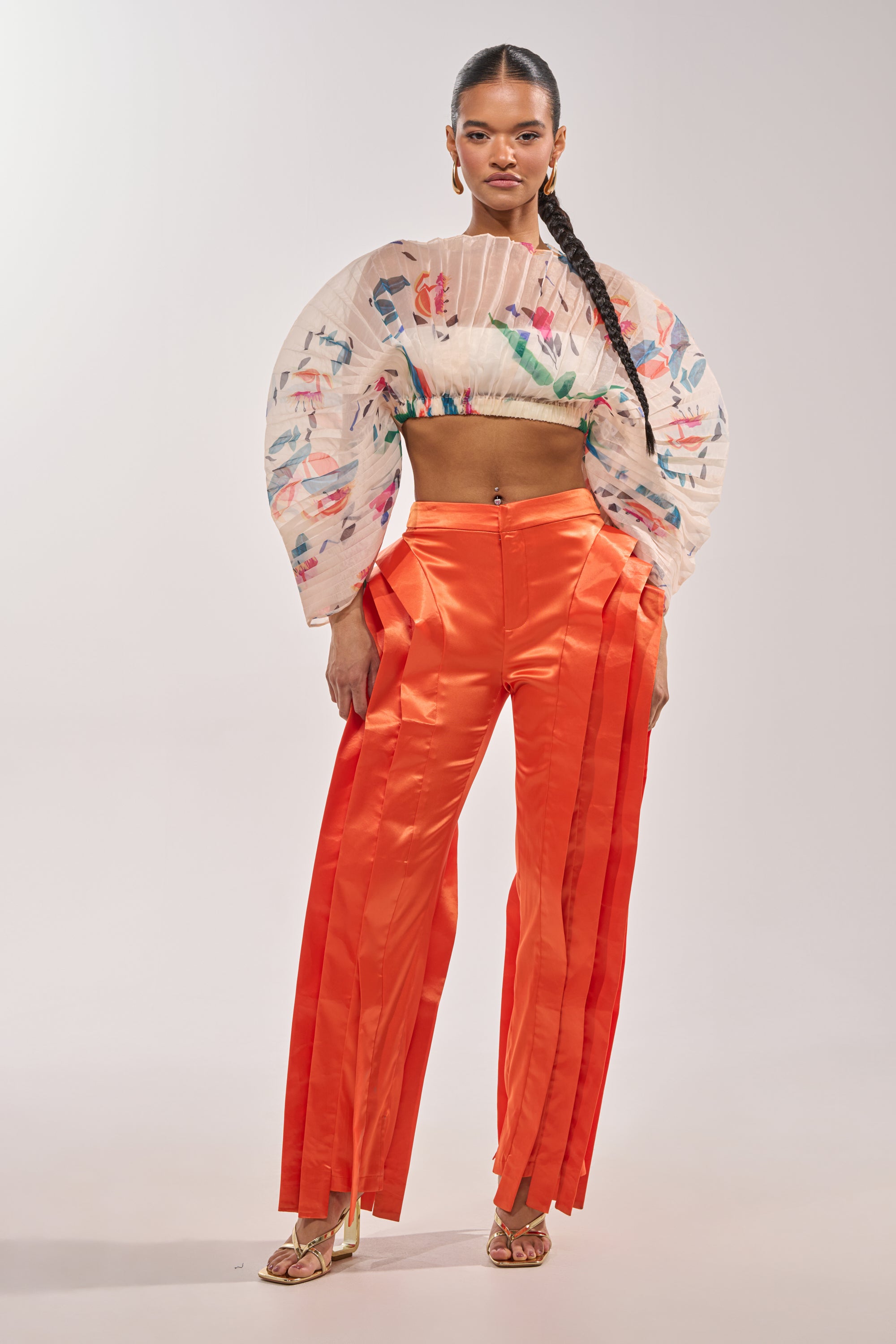 A woman poses confidently in the INFINITY TROUSER IN ORANGE, featuring a high-rise waist and bold satin finish, paired with a sheer floral crop top, gold heeled sandals, and long braided hair against a plain background. 1