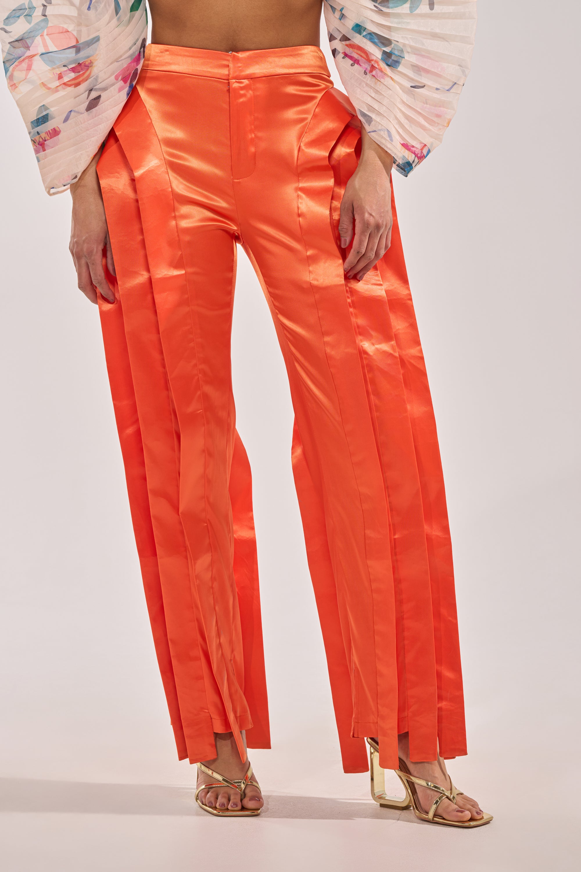Wearing the INFINITY TROUSER IN ORANGE—bright satin, high-rise waist, front pleats, slit hems—paired with gold strappy heels and a voluminous floral top. 0