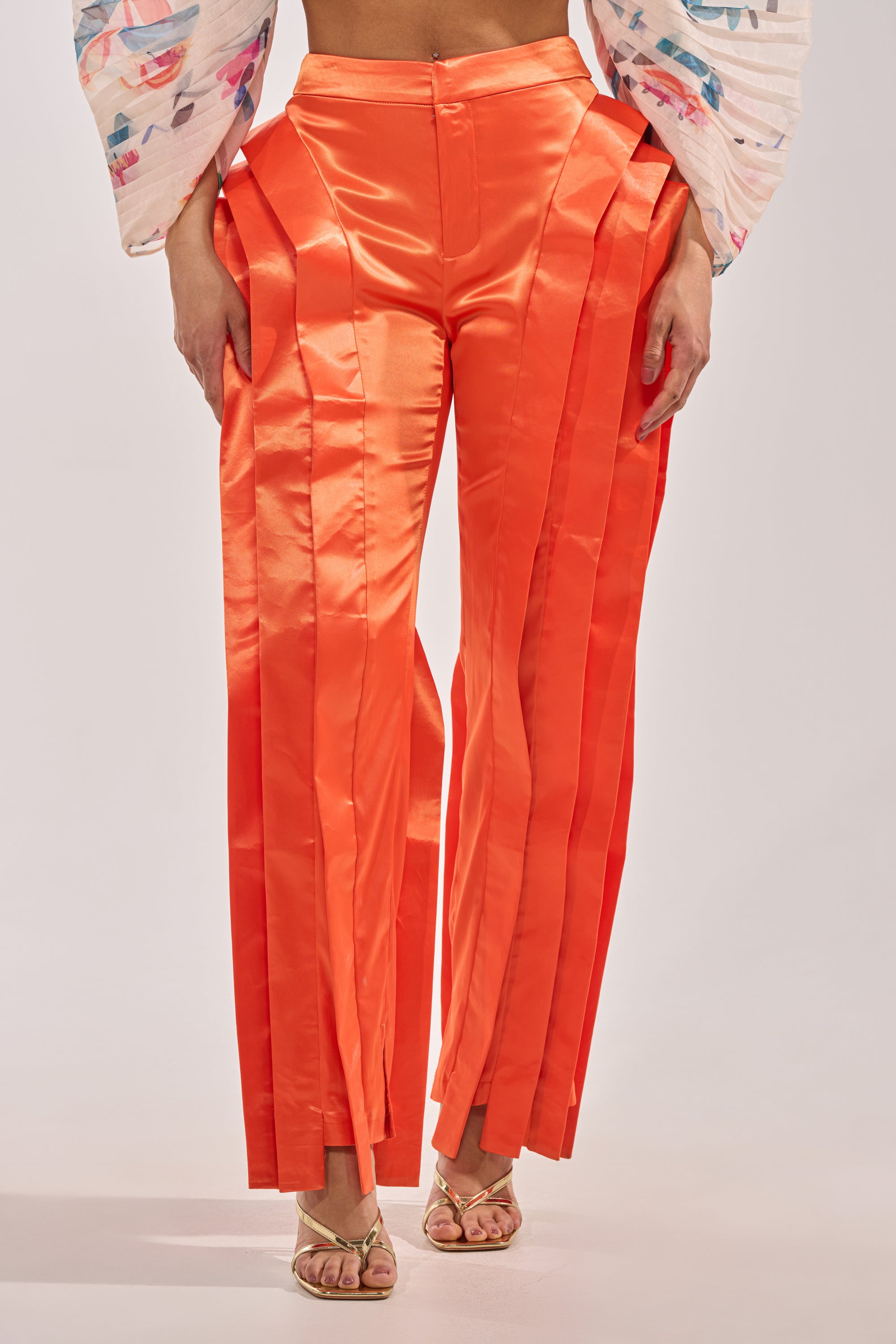 Wearing the INFINITY TROUSER IN ORANGE—vibrant satin statement pants with pleats and a high-rise waist—styled with a white patterned long-sleeve top and gold open-toe heels. 2