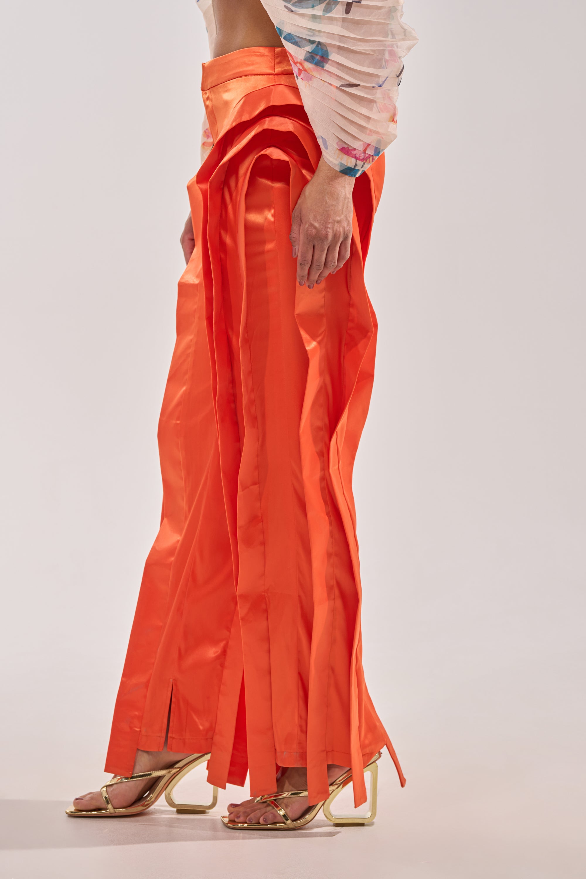 A person wearing the INFINITY TROUSER IN ORANGE—bright orange, satin, high-rise pants with ankle slits—styled with gold high-heeled sandals and a sheer floral long-sleeve top. The background is plain light gray. 3