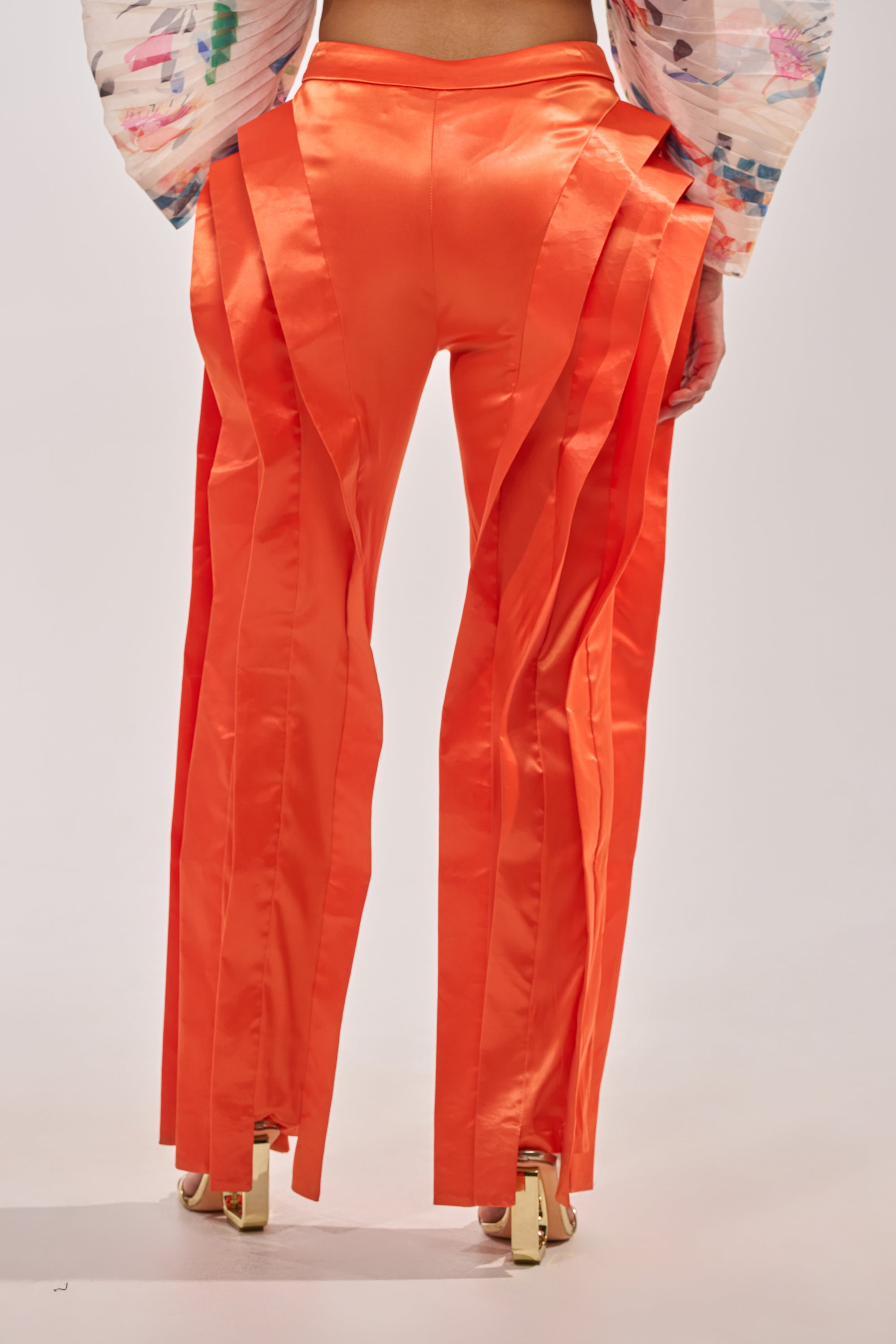 A person shown from the back wears the INFINITY TROUSER IN ORANGE—bright satin, high-rise trousers with layered vertical strips—paired with a colorful textured top and gold high-heeled sandals. 4