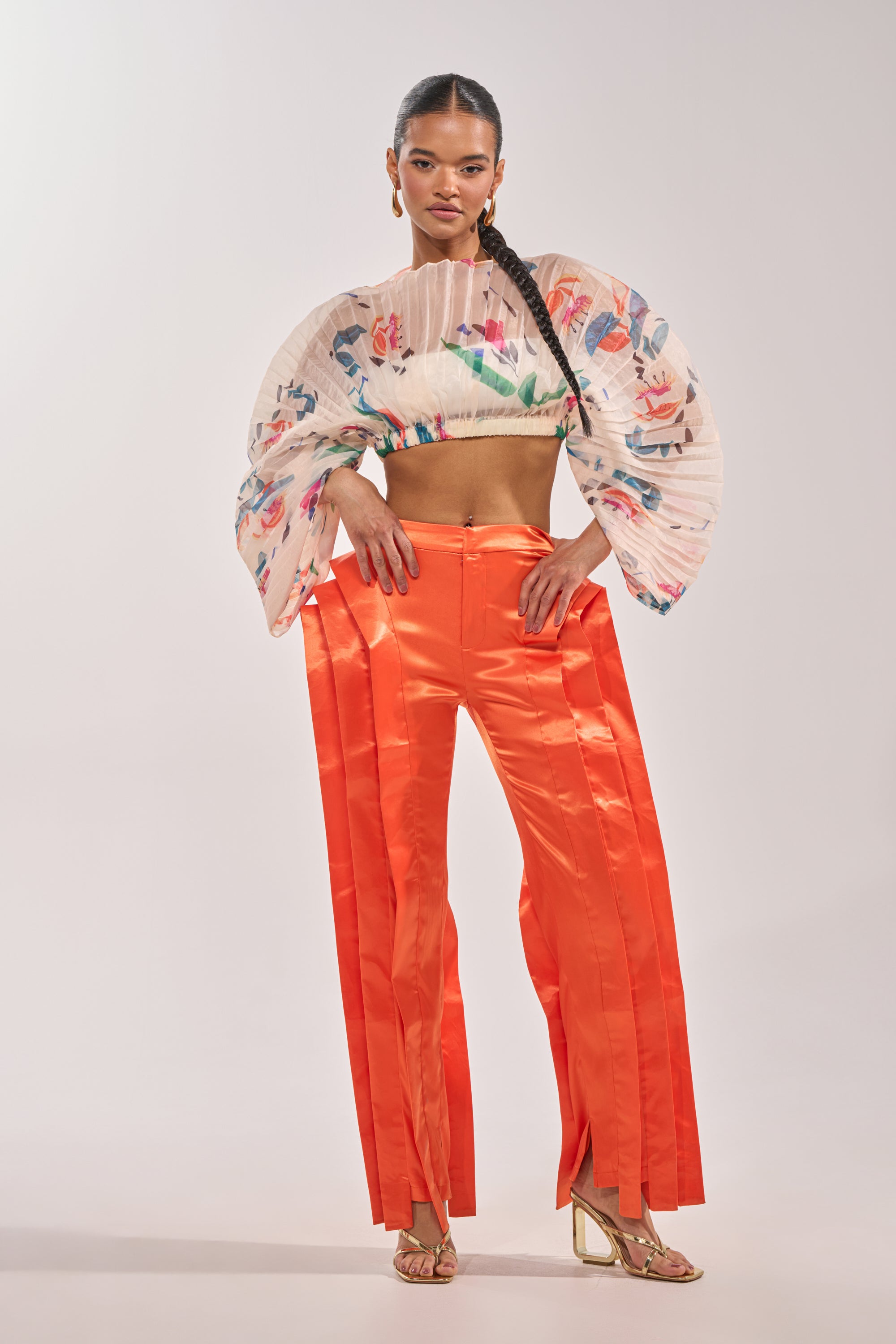 A woman poses in the INFINITY TROUSER IN ORANGE—shiny, satin, high-rise statement pants—paired with a sheer floral pleated crop top, gold open-toe heels, and her hair in a long braid. 5
