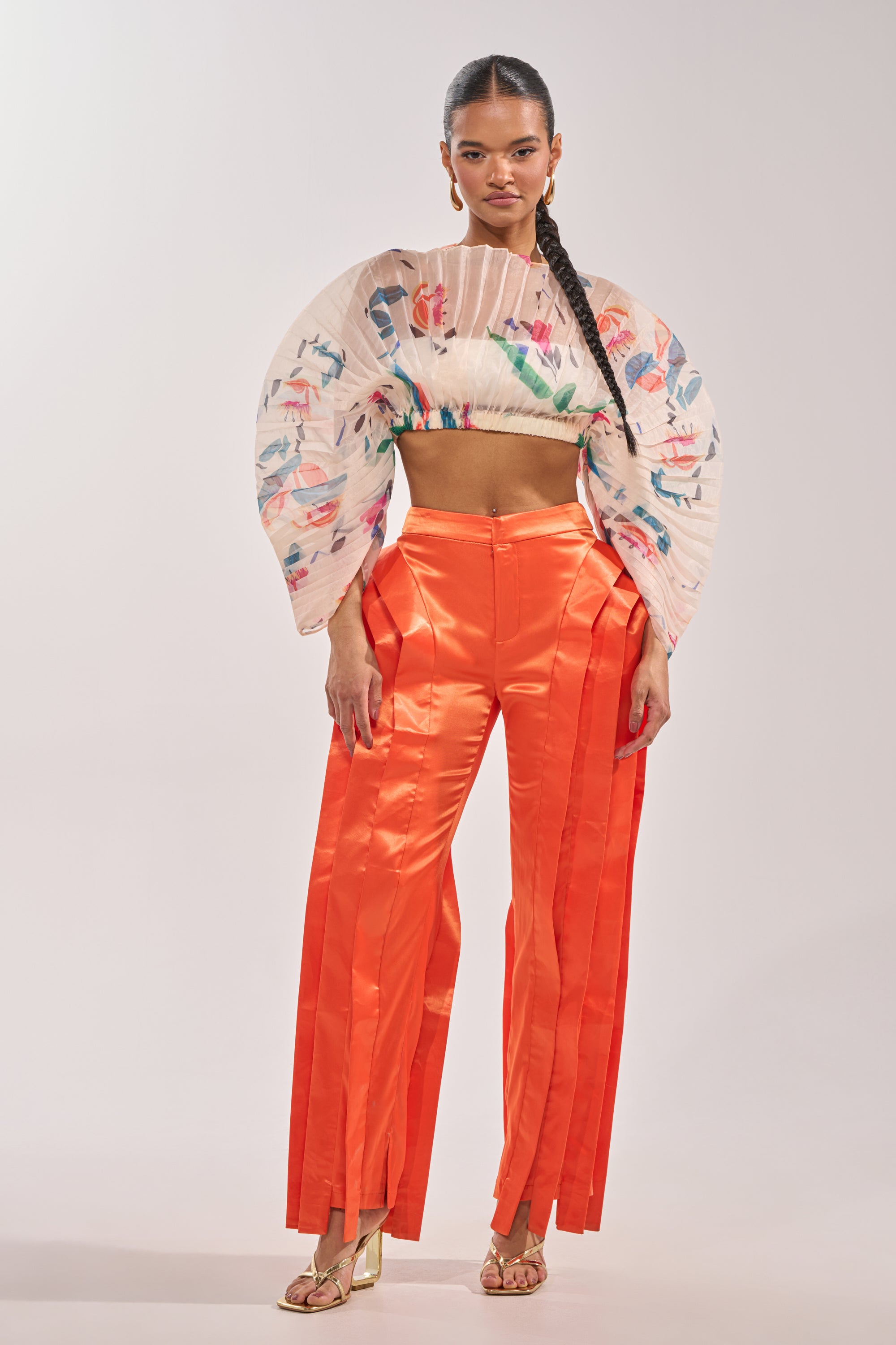 A woman faces forward, wearing a pleated floral crop top with dramatic sleeves and the INFINITY TROUSER IN ORANGE—satin statement pants featuring a high-rise waist—paired with gold heeled sandals and her hair in a long braid. 6