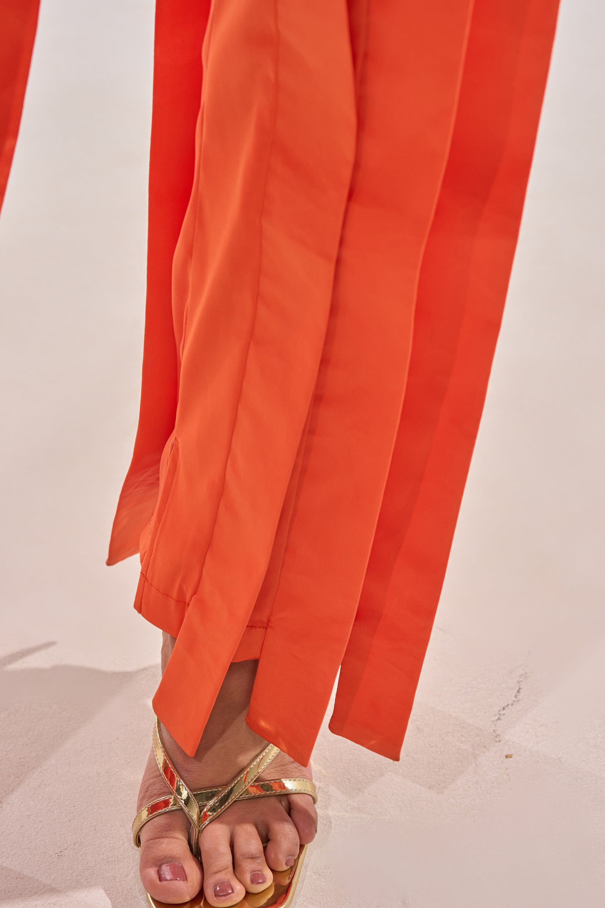 A close-up of a person wearing the INFINITY TROUSER IN ORANGE, featuring bright satin fabric, a high-rise waist, and hem slits, paired with gold sandals that reveal painted toenails on a light-colored surface. 7